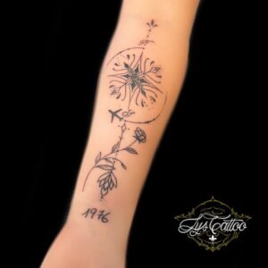 Tatouage boussole minimaliste sur l’avant-bras, agrémenté d’un avion, de fleurs et d’une date, réalisé par Lys Tattoo à Gradignan, près de Bordeaux et Pessac.