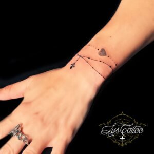 Tatouage fin et minimaliste sur le poignet, représentant une flèche entourant le poignet avec un cœur en pointillés, réalisé par Lys Tattoo à Gradignan (Bordeaux).