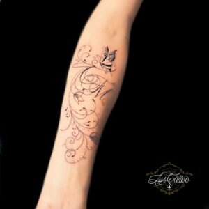 Tatouage fin et délicat sur l’avant-bras représentant des arabesques florales, la lettre "M" en calligraphie élégante et un papillon, réalisé par Lys Tattoo à Gradignan (Bordeaux).