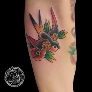 Tatouage d'hirondelle colorée de style traditionnel américain, entourée de fleurs vibrantes, réalisé sur l'avant-bras par Le Bull Tatoué au salon Lys Tattoo.