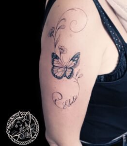 Tatouage délicat d’un papillon accompagné de fleurs et d’arabesques, avec le prénom "Élodie", réalisé par Le Bull Tatoué du salon Lys Tattoo à Gradignan, près de Bordeaux.