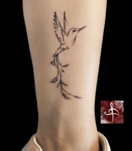 Petit colibri en fine line sur cheville, queue feuillue et points subtils – Rhum1 Tattoo Bordeaux