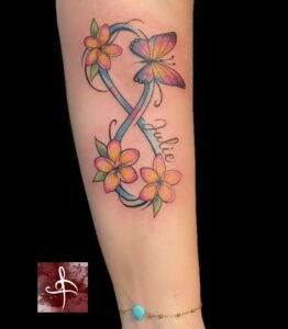 Tatouage signe infini coloré avec fleurs de frangipanier, papillon et prénom Julie – avant-bras (Rhum1 Tattoo)