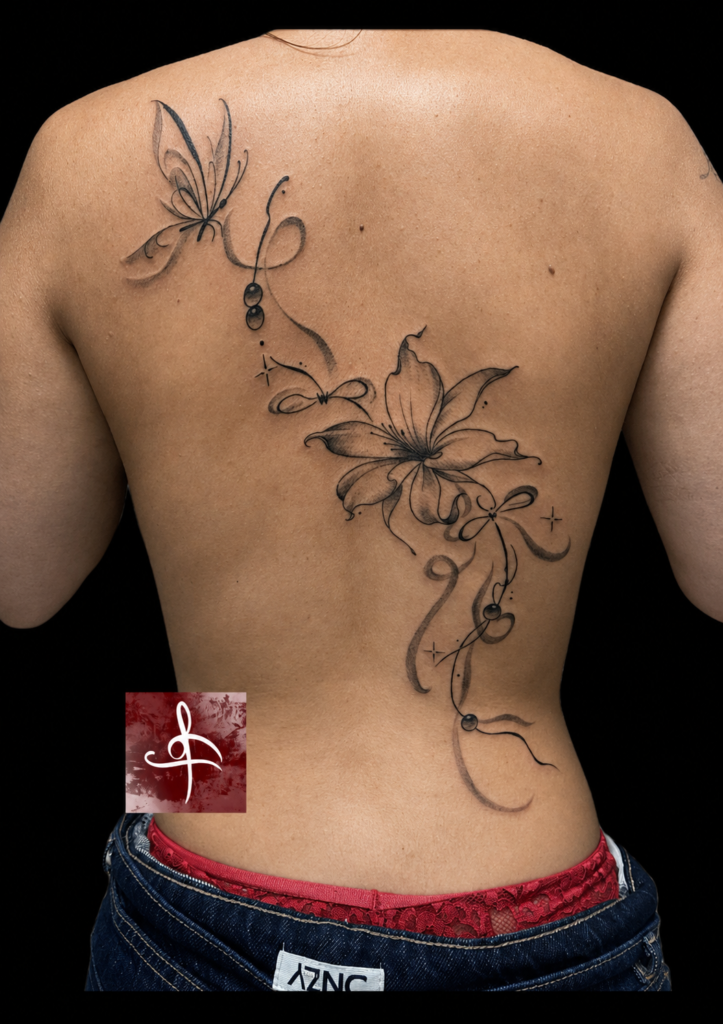 Tatouage floral dans le dos composé d’une grande fleur de lys, d’un papillon, de volutes et de perles en noir et gris, réalisé chez Lys Tattoo à Villenave-d’Ornon.
