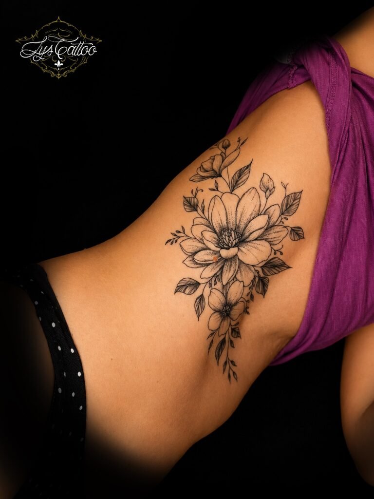 Tatouage floral noir et gris sur les côtes, composé d’une grande fleur de magnolia et de fleurs délicates, réalisé en style fineline chez Lys Tattoo à Villenave-d’Ornon.