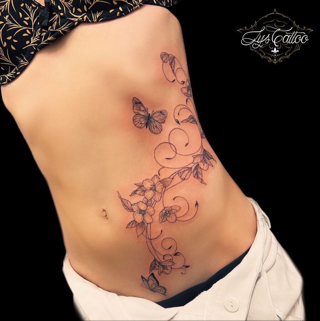 Tatouage floral fineline avec fleurs de cerisier, papillons et arabesques sur les côtes, réalisé chez Lys Tattoo à Villenave-d’Ornon près de Bordeaux.